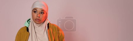 pensive multiracial woman in hijab and yellow stylish jacket looking away on pinkish grey background, banner
