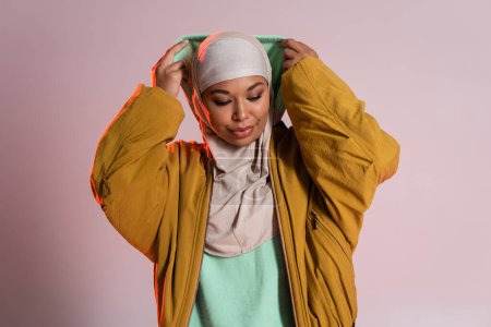 young and trendy multiracial woman in hijab and yellow bomber jacket wearing hood on pinkish grey background