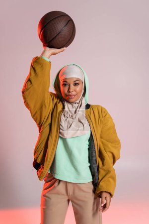 mujer multirracial con estilo en hijab y chaqueta de bombardero amarillo de pie con baloncesto en la mano levantada sobre fondo gris y rosa