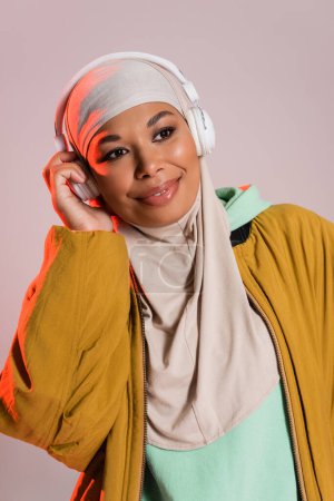 Unbekümmerte multirassische muslimische Frau in gelber Jacke, die in drahtlosen Kopfhörern isoliert auf grau Musik hört