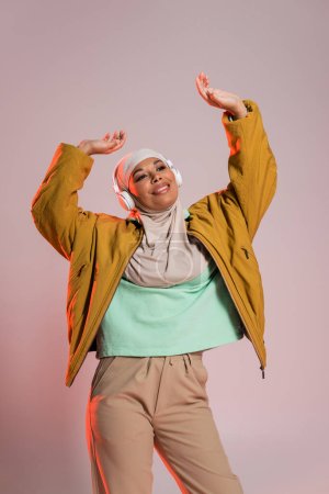 joyful multiracial muslim woman in stylish casual clothes and headphones dancing with raised hands on pinkish grey background