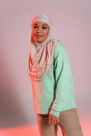 pretty multiracial woman in traditional hijab and green long sleeve shirt smiling at camera on grey and pink background