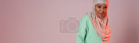 Photo for Positive and pretty multiracial woman in traditional muslim hijab smiling at camera on pinkish grey background, banner - Royalty Free Image