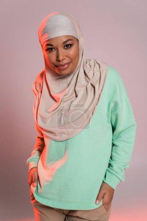 fashionable multiracial woman in hijab and green long sleeve shirt smiling at camera on pinkish grey background