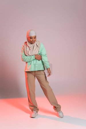 pleine longueur de jeune femme multiraciale en hijab et pantalon beige avec des baskets debout sur fond gris et rose