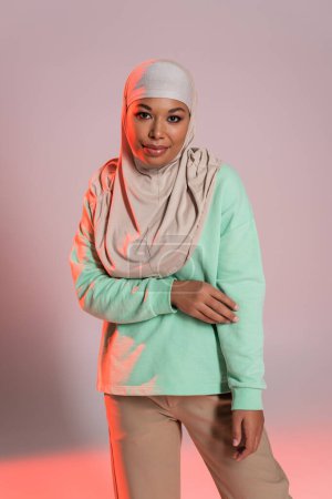pretty multiracial woman in muslim hijab and green long sleeve shirt looking at camera on grey and pink background