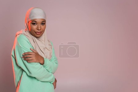 young multiracial muslim woman in traditional hijab and green long sleeve shirt looking at camera while posing on pinkish grey