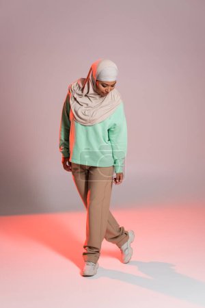 volle Länge der multirassischen Frau im Hijab und langärmeligem Hemd mit Hose auf grauem und rosa Hintergrund