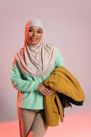 cheerful multiracial muslim woman in hijab holding yellow jacket and smiling at camera on grey and pink background