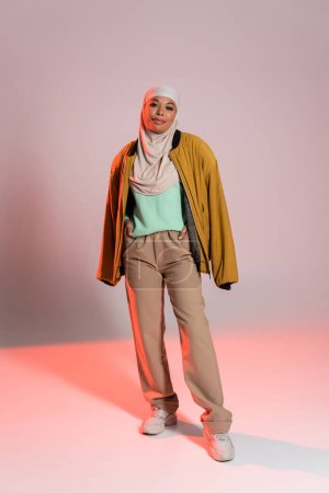 full length of multiracial muslim woman in yellow jacket and beige pants with sneakers on grey and pink background