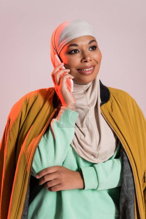 carefree multiracial woman in hijab and yellow jacket talking on cellphone and looking away isolated on grey