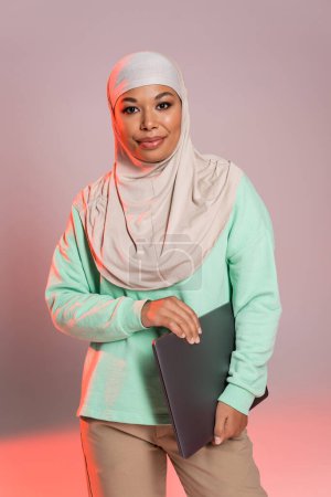 happy multiracial woman in hijab holding laptop and smiling at camera on grey and pink background