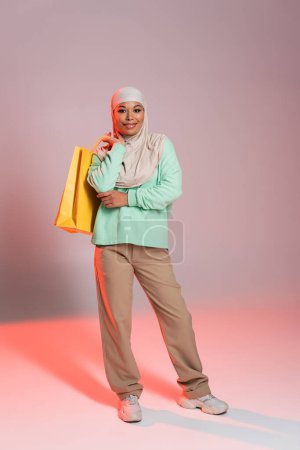 pleine longueur de femme multiraciale heureuse en hijab et tenue décontractée tendance posant avec des sacs à provisions jaunes sur fond gris et rose