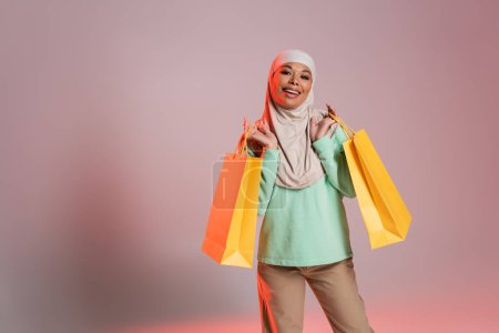 fröhliche multirassische Frau in trendiger Freizeitkleidung und traditionellem Hijab mit gelben Einkaufstaschen auf rosa-grauem Hintergrund