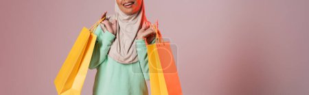 abgeschnittene Ansicht einer glücklichen multirassischen muslimischen Frau mit gelben Einkaufstaschen auf rosa grauem Hintergrund, Banner