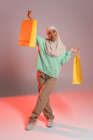 in voller Länge fröhliche multirassische Frau im muslimischen Hijab posiert mit gelben Einkaufstaschen auf grauem und pinkem Hintergrund