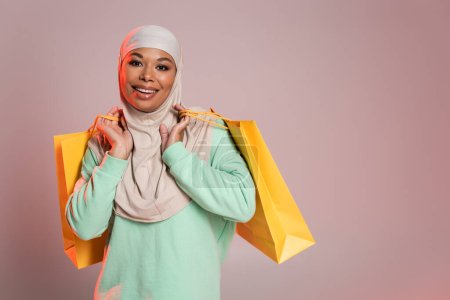 overjoyed multiracial woman in muslim hijab holding yellow shopping bags and smiling at camera on pinkish grey