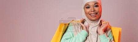 carefree multiracial woman in hijab posing with yellow shopping bags and smiling at camera isolated on grey, banner