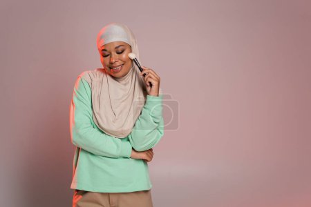 pleased multiracial muslim woman in hijab holding cosmetic brush and smiling with closed eyes on pinkish grey background