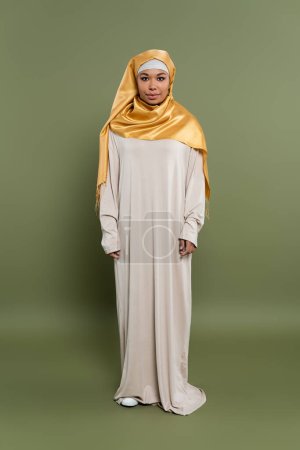 full length of multiracial muslim woman in yellow silk hijab and abaya dress standing on olive green background