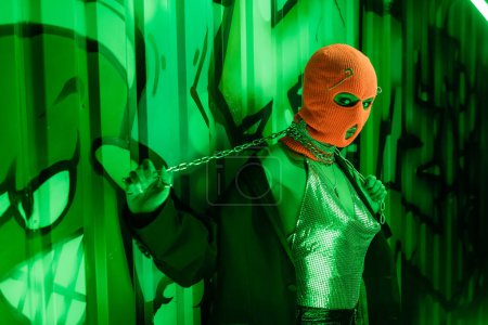 provocative woman in balaclava and sexy outfit posing with neck chain near wall with graffiti in green lighting