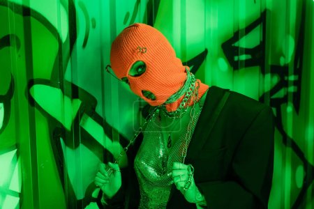 Photo for Seductive woman in orange balaclava and black blazer with silver top posing with metallic chain near green wall with graffiti - Royalty Free Image