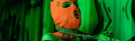 Photo for Seductive woman in orange balaclava holding metallic neck chains near green wall with graffiti, banner - Royalty Free Image