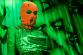 low angle view of young woman in balaclava and sexy top looking at camera in green lighting near wall with graffiti Poster #645512990