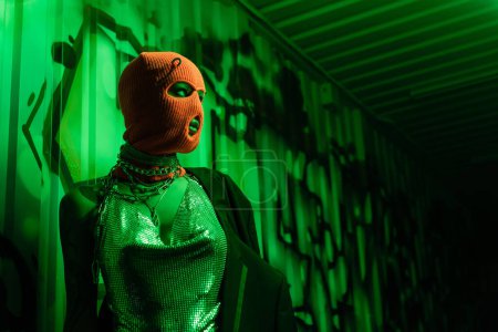 Photo for Anonymous sexy woman in orange balaclava and silver top looking away near wall with graffiti in green light - Royalty Free Image
