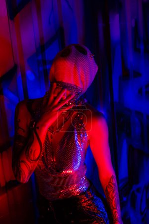 Photo for Passionate tattooed woman in balaclava and silver top touching neck chains near blue wall with graffiti in red light - Royalty Free Image