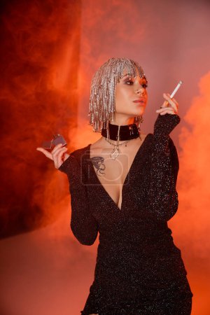 Photo for Extravagant tattooed woman in silver wig and sexy dress posing with cigarette on red and orange background with smoke - Royalty Free Image