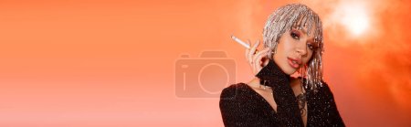 Porträt einer sinnlich tätowierten Frau in metallischer Kopfbedeckung mit Zigarette auf korallenrosa rauchigem Hintergrund, Banner
