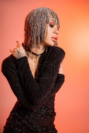 Téléchargez les photos : Séduisante femme à la mode en bijoux coiffure et robe lurex noir s'embrassant tout en posant sur fond rose corail - en image libre de droit