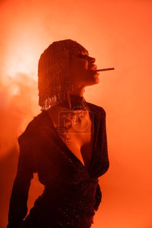 Foto de Seductora mujer tatuada en peluca metálica y vestido sexy negro posando con cigarrillo sobre fondo rojo y naranja - Imagen libre de derechos