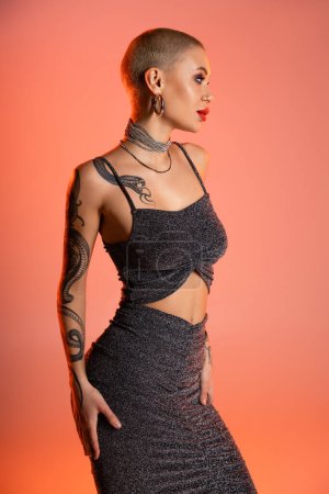 tattooed woman in grey lurex crop top and skirt looking away on coral pink background