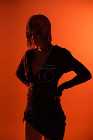 silhouette of woman in black lurex dress and metallic headwear standing with hands on waist on orange background