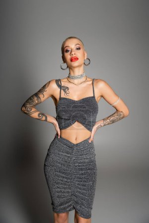 extravagant tattooed woman with short hair and bright makeup looking at camera while posing with hands on waist on grey background