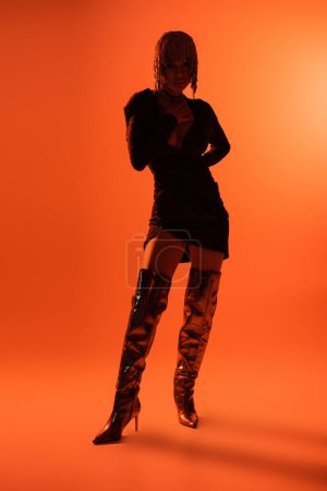 full length of seductive woman in long shiny boots and black dress standing with hand on waist on orange background
