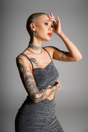 Photo for Tattooed woman with short hair and red lips holding hand near face and posing in glamour clothes isolated on grey - Royalty Free Image