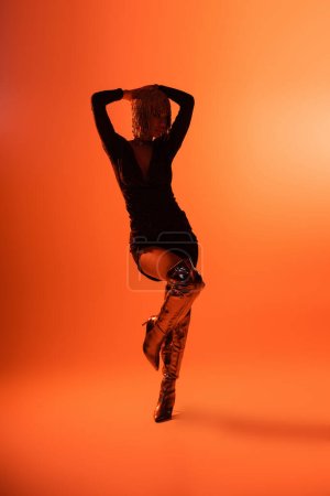 silhouette of woman in black dress and trendy long boots standing with hands above head on orange background