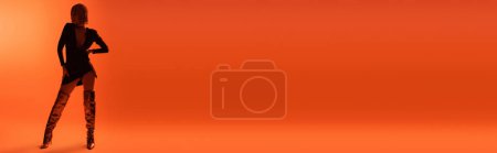 Photo for Full length of shaded woman in black dress and long boots standing with hand on hip on orange background, banner - Royalty Free Image