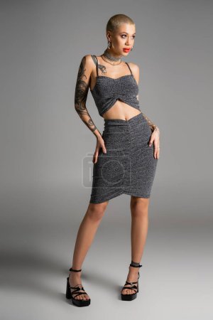 Photo for Full length of sexy tattooed woman in lurex skirt and crop top standing with hands on hips and looking away on grey background - Royalty Free Image