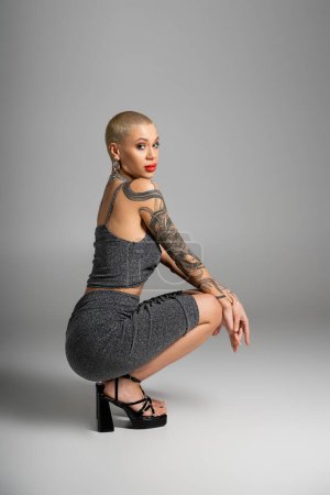 full length of sexy tattooed woman in lurex skirt and heeled sandals sitting on haunches on grey background