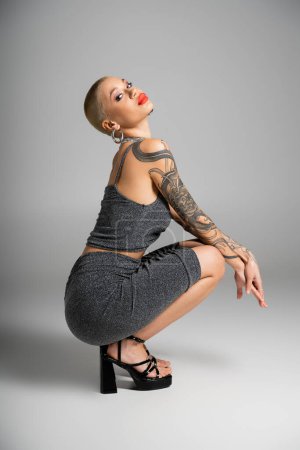 full length of tattooed and sexy woman in lurex clothes and sandals sitting on haunches and looking at camera on grey background
