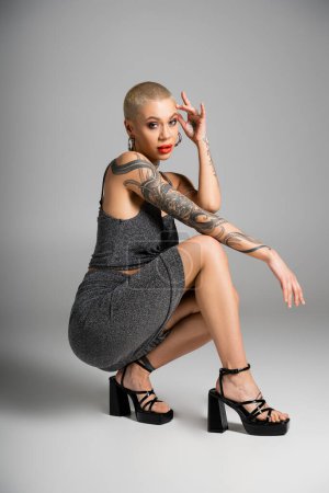 Photo for Full length of fashionable tattooed woman on heels sitting on haunches and looking at camera on grey background - Royalty Free Image