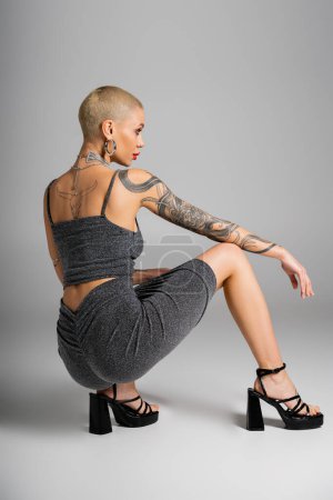 longitud completa de glamour mujer tatuada en traje sexy posando en las horquillas y mirando hacia otro lado sobre fondo gris