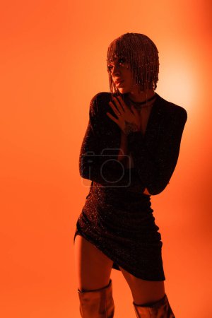sensual woman in black lurex dress and silver wig looking away while standing on orange background