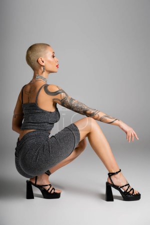 Photo for Side view of tattooed glamour woman in sexy outfit sitting on haunches on grey background - Royalty Free Image