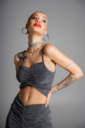 Téléchargez les photos : Femme tatouée extravagante avec des boucles d'oreilles cerceau et lèvres rouges posant avec la main à la taille et regardant loin isolé sur gris - en image libre de droit
