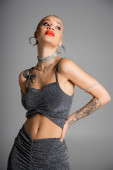 extravagant tattooed woman with hoop earrings and red lips posing with hand on waist and looking away isolated on grey hoodie #645513586